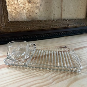 May include: A clear glass snack set consisting of a rectangular tray with a ribbed surface and a beaded edge, featuring a matching small cup with a beaded handle. The cup is designed to sit securely within a circular indentation on the tray.