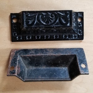 Puede incluir: Dos tiradores de cajón de metal negro. El tirador superior tiene un diseño adornado con un motivo floral. El tirador inferior es de forma rectangular simple.