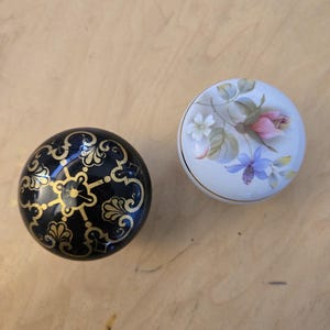 May include: Two round ceramic cabinet knobs. One knob is black with a gold-coloured filigree pattern. The other knob is white with a hand-painted floral design featuring a pink rose, blue flowers, green leaves, and a thin gold-coloured rim.