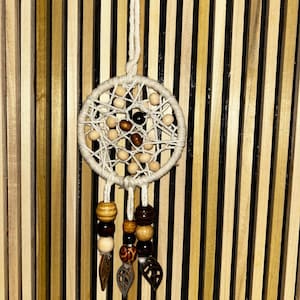 May include: A handmade dreamcatcher with a circular woven web design, featuring wooden beads in various shades of brown and cream. The dreamcatcher hangs from a white string and has decorative beaded strands with leaf charms.