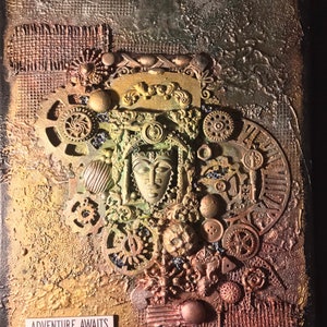 May include: A mixed media art piece with a gold, green, and brown color scheme. The piece features a central image of a face with intricate details and a variety of gears, buttons, and other embellishments. The text "Adventure Awaits" is printed at the bottom of the piece.