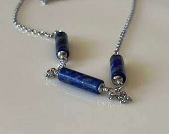 Op de afbeelding: Een zilveren ketting met drie blauwe lapis lazuli kralen. De kralen zijn cilindrisch en hebben een marmerpatroon.