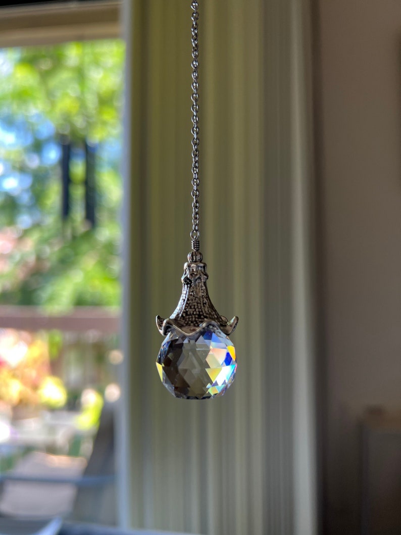 Op de afbeelding: Een gefacetteerde kristallen bol hanger hangt aan een zilveren ketting. Het kristal reflecteert een regenboog aan kleuren. De hanger heeft een decoratieve zilveren dop met een textuur. De achtergrond is wazig.