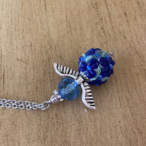 Op de afbeelding: Een zilveren engel hanger ketting met een blauwe glazen kraal. De kraal is versierd met witte en blauwe bloemen.
