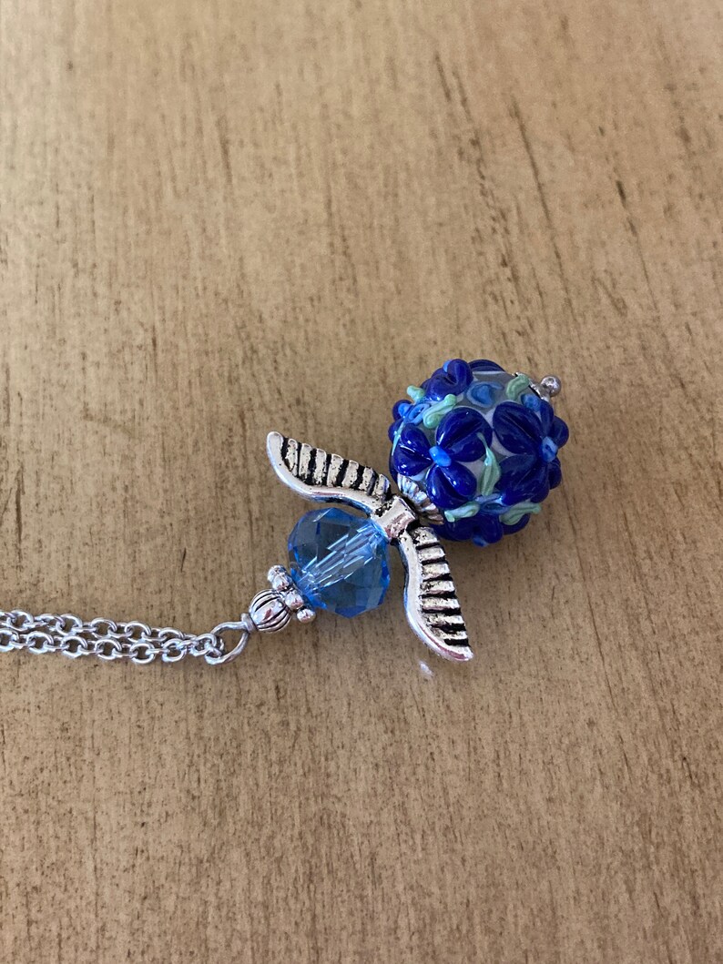 Op de afbeelding: Een zilveren engel hanger ketting met een blauwe glazen kraal. De kraal is versierd met witte en blauwe bloemen.