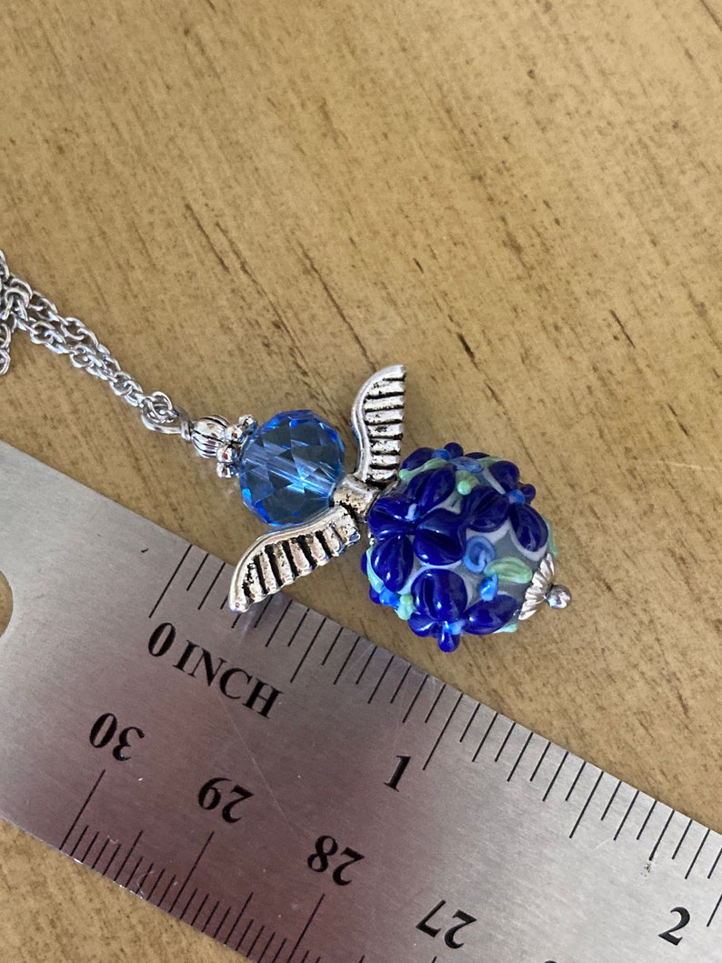 Op de afbeelding: Een zilveren ketting met een blauwe glazen kraal en een zilveren engelenvleugel bedel. De kraal is ongeveer 2,5 cm in diameter.