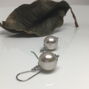 May include: A pair of pearl earrings with a silver-colored hook. The earrings feature large, round, light-colored pearls with a silver-colored decorative cap. A dried, dark green leaf is in the background.