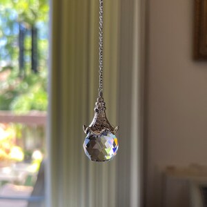 Op de afbeelding: Een gefacetteerde kristallen bol zonnevanger hangend aan een zilveren ketting. Het kristal reflecteert een regenboog aan kleuren. De zonnevanger hangt voor een vage achtergrond van een raam en interieur.