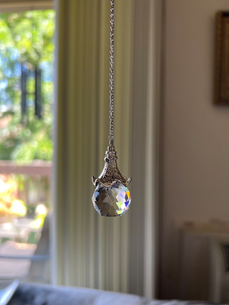 Op de afbeelding: Een gefacetteerde kristallen bol zonnevanger hangend aan een zilveren ketting. Het kristal reflecteert een regenboog aan kleuren. De zonnevanger hangt voor een vage achtergrond van een raam en interieur.