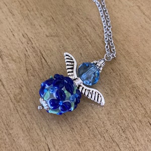 Op de afbeelding: Een zilveren engelhangerketting met een blauwe glazen kraal met een bloemenmotief. De kraal is omgeven door zilveren vleugels en een blauwe kristal.
