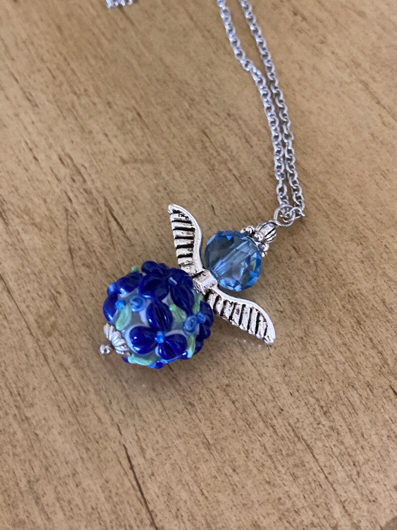 Op de afbeelding: Een zilveren engelhangerketting met een blauwe glazen kraal met een bloemenmotief. De kraal is omgeven door zilveren vleugels en een blauwe kristal.
