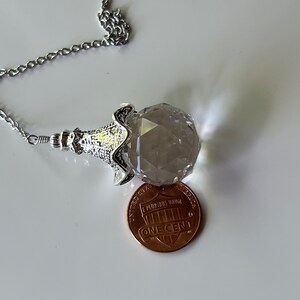 Op de afbeelding: Een gefacetteerde kristallen hangerketting met een zilverkleurige ketting. De kristal wordt vastgehouden in een decoratieve zilverkleurige setting. Een munt ligt naast de kristal ter vergelijking.