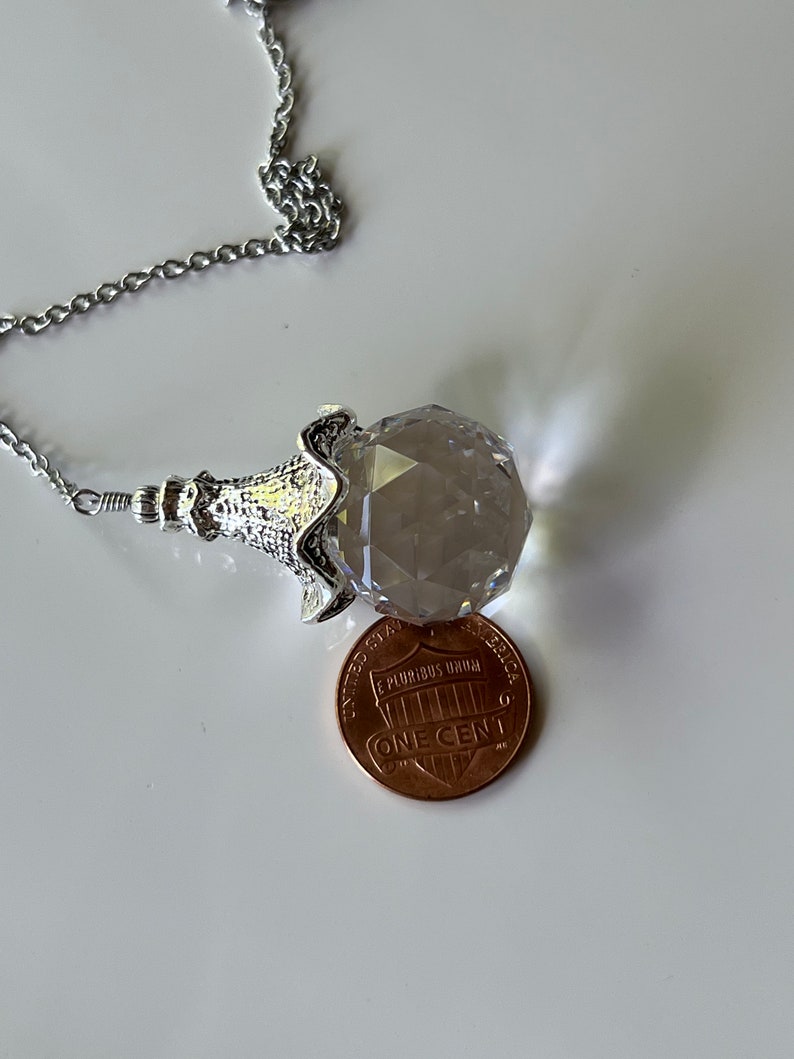 Op de afbeelding: Een gefacetteerde kristallen hangerketting met een zilverkleurige ketting. De kristal wordt vastgehouden in een decoratieve zilverkleurige setting. Een munt ligt naast de kristal ter vergelijking.