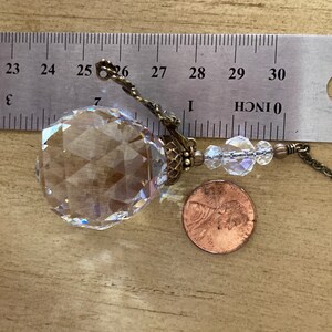 May include: A clear crystal ball with a faceted design, hanging from a brass chain with a small brass clasp. The crystal ball is about 1 inch in diameter and is next to a US penny for scale.