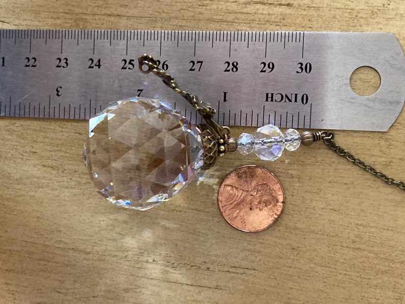 May include: A clear crystal ball with a faceted design, hanging from a brass chain with a small brass clasp. The crystal ball is about 1 inch in diameter and is next to a US penny for scale.