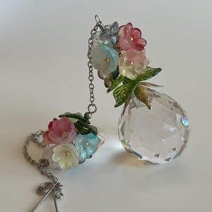May include: A clear crystal prism with a teardrop shape, adorned with colorful floral accents. The prism is connected by a silver chain to a cluster of matching flowers. The flowers are in shades of pink, blue, and yellow, with green leaves.
