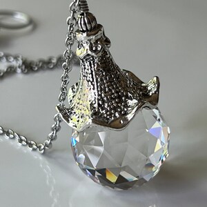 Op de afbeelding: Een heldere, facetgeslepen kristallen bolhanger met een zilverkleurige metalen dop en ketting. Het kristal reflecteert licht en cre&euml;ert een regenboogeffect. De hanger hangt aan een delicate ketting, met een klassiek sieradenontwerp.