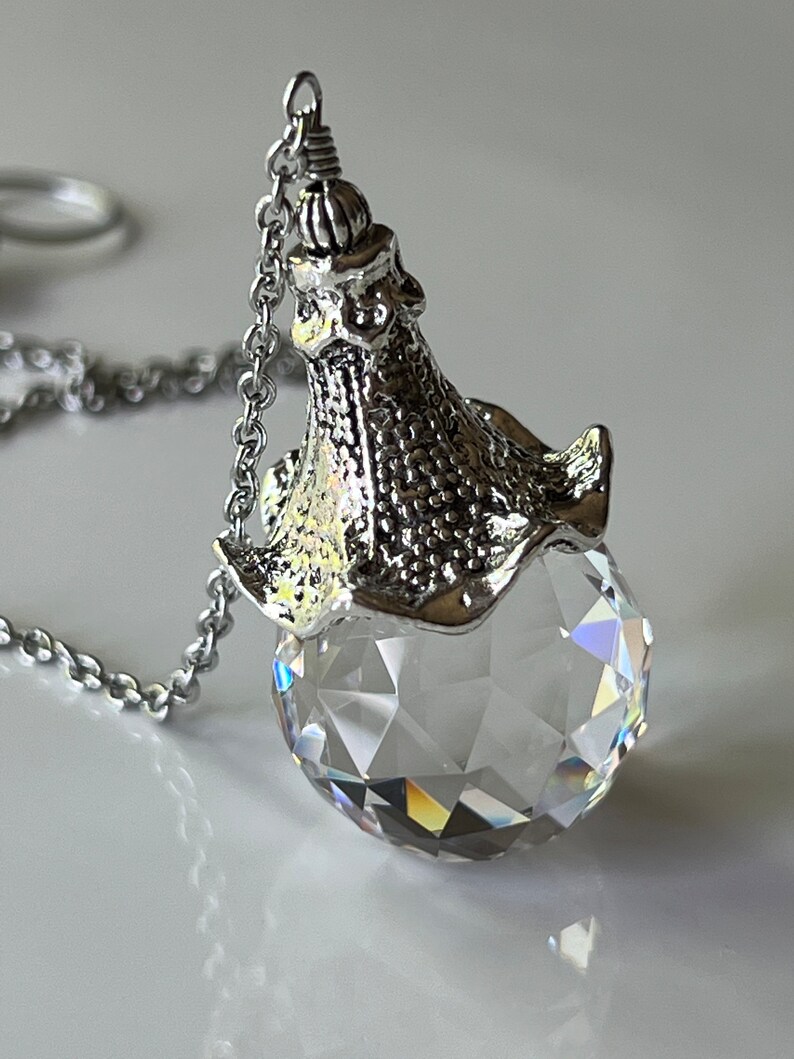 Op de afbeelding: Een heldere, facetgeslepen kristallen bolhanger met een zilverkleurige metalen dop en ketting. Het kristal reflecteert licht en cre&euml;ert een regenboogeffect. De hanger hangt aan een delicate ketting, met een klassiek sieradenontwerp.