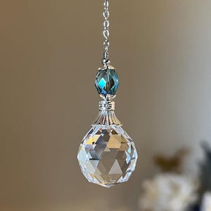 May include: A clear, faceted crystal ball pendant hangs from a silver chain. Above the ball is a blue-green faceted bead and decorative silver accents. The pendant is suspended against a soft, blurred background.