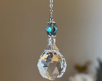 May include: A clear, faceted crystal ball pendant hangs from a silver chain. Above the ball is a blue-green faceted bead and decorative silver accents. The pendant is suspended against a soft, blurred background.
