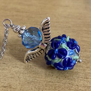 Op de afbeelding: Een zilveren ketting met een bedel, een blauwe kristal en een blauwe en witte glazen kraal met een bloemenpatroon.