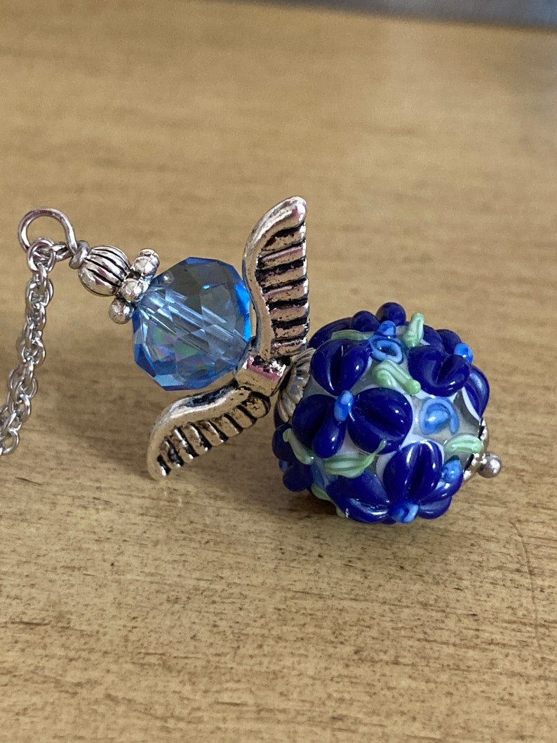Op de afbeelding: Een zilveren ketting met een bedel, een blauwe kristal en een blauwe en witte glazen kraal met een bloemenpatroon.