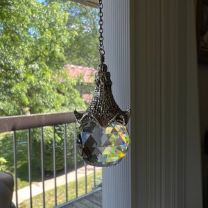 Op de afbeelding: Een heldere kristallen bol zonnevanger hangend aan een zilveren ketting. Het kristal is facetgeslepen en reflecteert een regenboog aan kleuren. De decoratieve metalen dop heeft een sierlijk ontwerp.