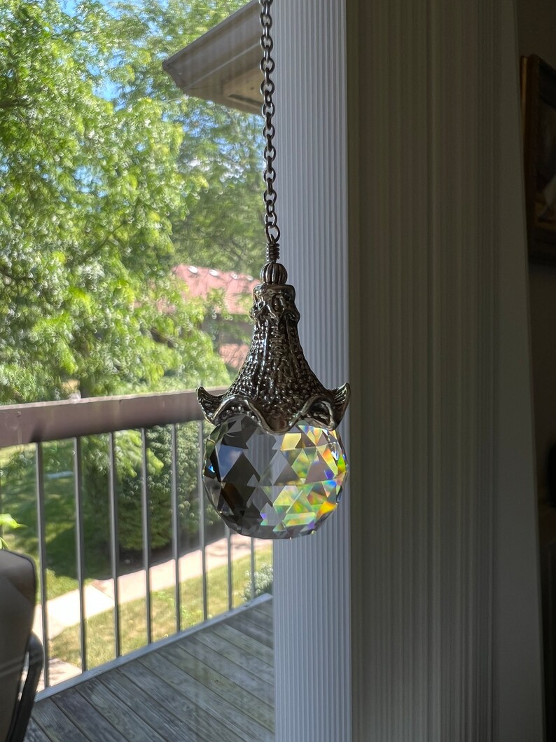 Op de afbeelding: Een heldere kristallen bol zonnevanger hangend aan een zilveren ketting. Het kristal is facetgeslepen en reflecteert een regenboog aan kleuren. De decoratieve metalen dop heeft een sierlijk ontwerp.