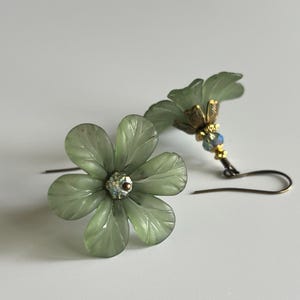 May include: A pair of delicate flower earrings. Each earring features a light green, five-petal flower with a sparkling center. The earrings have gold-colored accents and a hook closure. The earrings are a floral accessory.