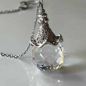 Op de afbeelding: Een zilverkleurige ketting met een facetgeslepen kristallen bolhanger. De hanger hangt aan een fijne ketting en heeft een decoratieve, getextureerde dop. De kristallen bol reflecteert licht en cre&euml;ert een sprankelend effect.