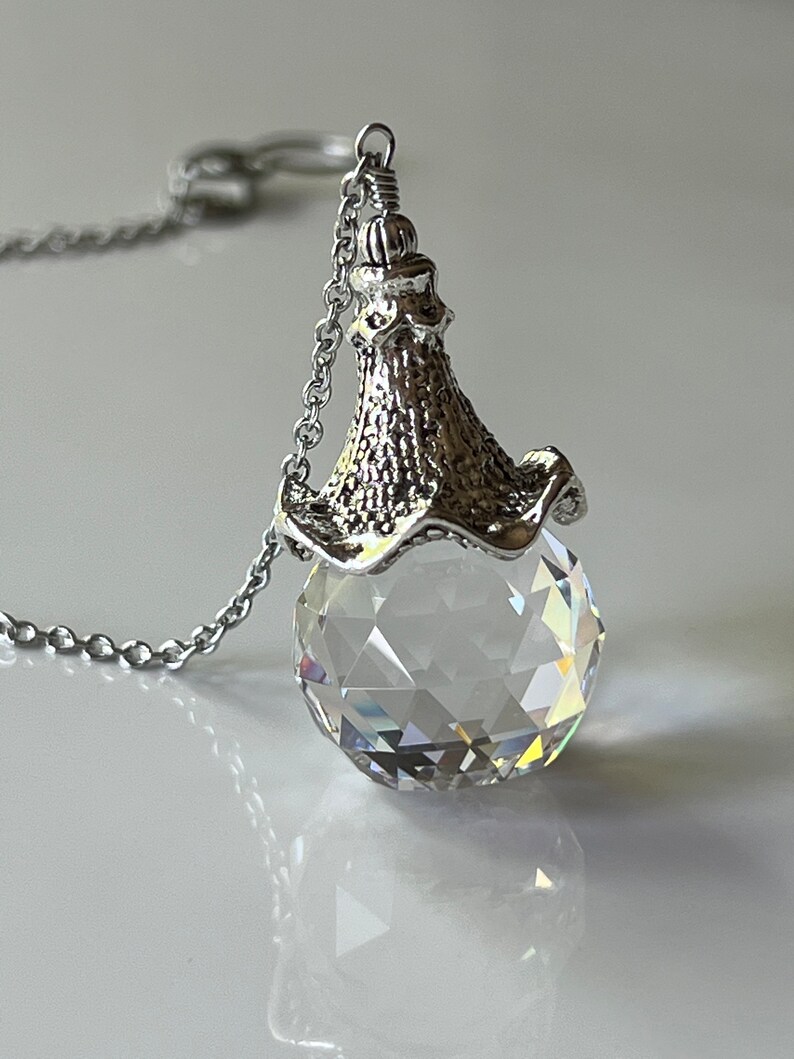 Op de afbeelding: Een zilverkleurige ketting met een facetgeslepen kristallen bolhanger. De hanger hangt aan een fijne ketting en heeft een decoratieve, getextureerde dop. De kristallen bol reflecteert licht en cre&euml;ert een sprankelend effect.