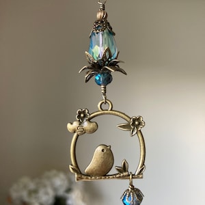 May include: A bronze-colored charm with a bird perched on a branch inside a circular frame. The frame is decorated with flowers and a cloud. The charm is attached to a chain with a blue crystal teardrop at the top and a smaller blue crystal teardrop at the bottom.