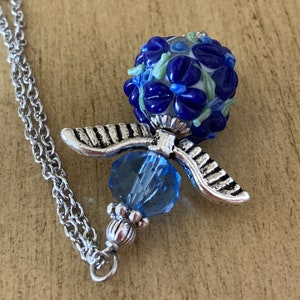 Op de afbeelding: Een zilveren ketting met een blauwe glazen kralenhanger. De kraal is versierd met blauwe en witte bloemen en heeft een zilveren engelenvleugelbedel.