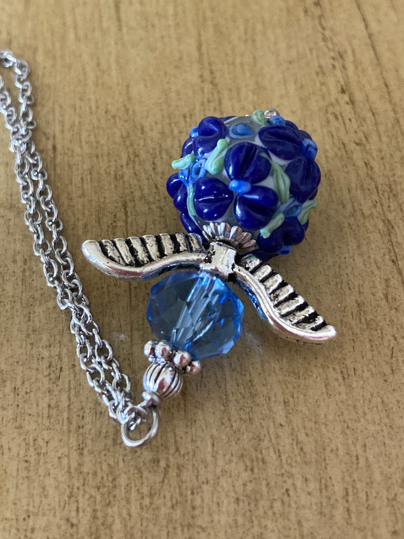 Op de afbeelding: Een zilveren ketting met een blauwe glazen kralenhanger. De kraal is versierd met blauwe en witte bloemen en heeft een zilveren engelenvleugelbedel.