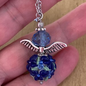 Op de afbeelding: Een zilveren ketting met een blauwe kristallen kraal en een zilveren engelenvleugelbedel. De bedel is bevestigd aan een blauwe en witte glazen kraal met een bloemenpatroon.