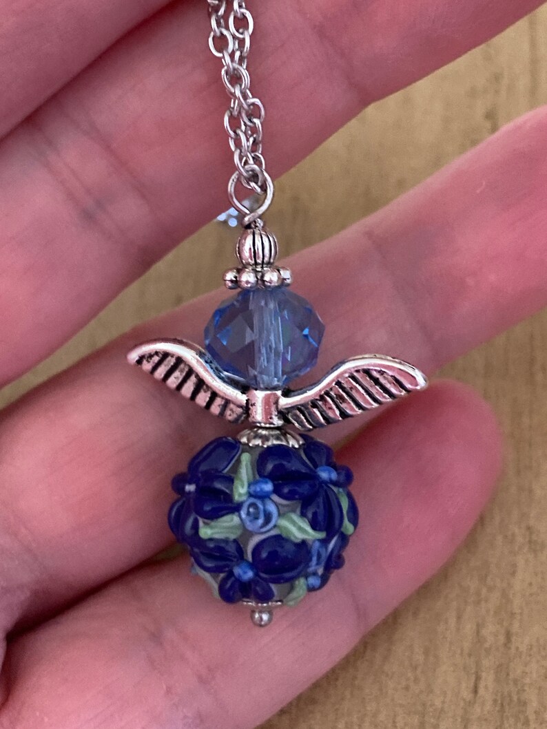 Op de afbeelding: Een zilveren ketting met een blauwe kristallen kraal en een zilveren engelenvleugelbedel. De bedel is bevestigd aan een blauwe en witte glazen kraal met een bloemenpatroon.