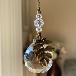 May include: A crystal suncatcher with a bronze leaf charm and a chain of clear glass beads. The suncatcher is hanging from a white ceiling.