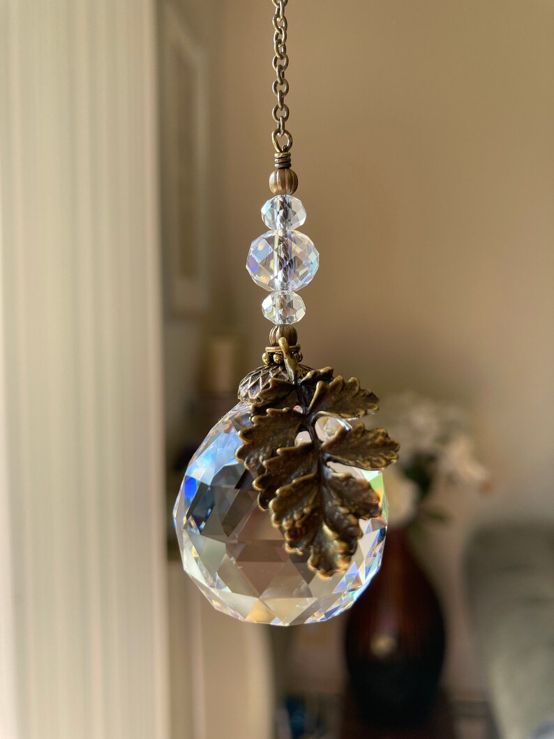May include: A crystal suncatcher with a bronze leaf charm and a chain of clear glass beads. The suncatcher is hanging from a white ceiling.