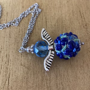 Op de afbeelding: Een zilveren engelenvleugel hangerketting met een blauwe glazen kraal met een bloemenmotief. De kraal is versierd met blauwe, witte en groene bloemen.
