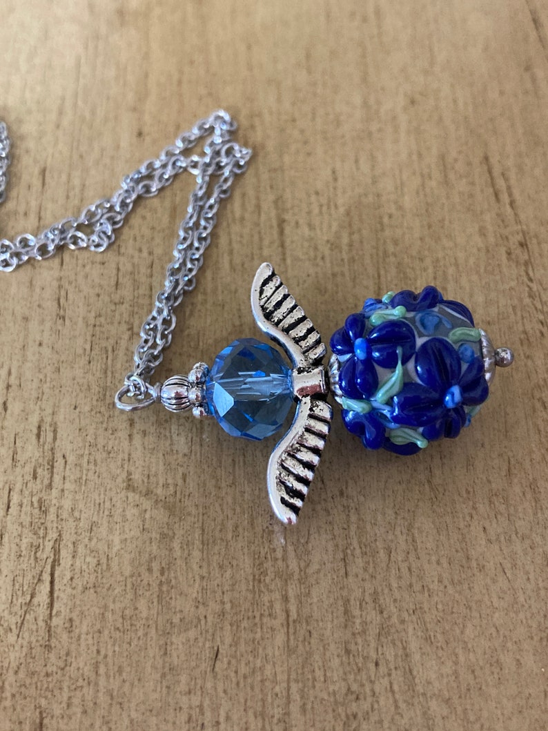 Op de afbeelding: Een zilveren engelenvleugel hangerketting met een blauwe glazen kraal met een bloemenmotief. De kraal is versierd met blauwe, witte en groene bloemen.