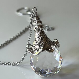 Op de afbeelding: Een zilverkleurige ketting met een facetgeslepen kristallen hanger. De heldere, bolvormige hanger heeft meerdere reflecterende oppervlakken, opgehangen aan een decoratieve metalen dop en ketting. De ketting is delicaat en de hanger schittert.