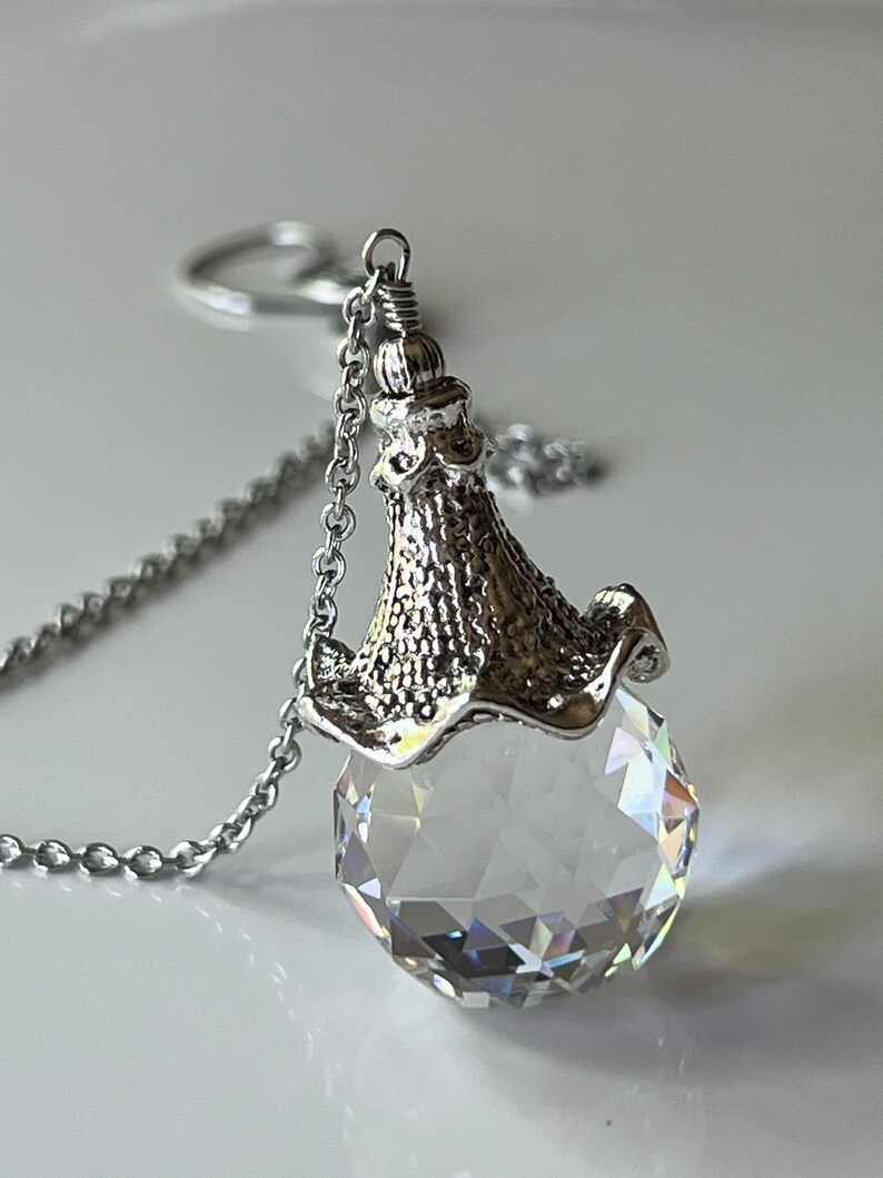 Op de afbeelding: Een zilverkleurige ketting met een facetgeslepen kristallen hanger. De heldere, bolvormige hanger heeft meerdere reflecterende oppervlakken, opgehangen aan een decoratieve metalen dop en ketting. De ketting is delicaat en de hanger schittert.