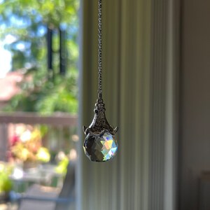 Op de afbeelding: Een gefacetteerde kristallen bol hanger hangt aan een zilveren ketting. Het kristal reflecteert een regenboog aan kleuren. De hanger hangt voor een raam met een vage buitenweergave.