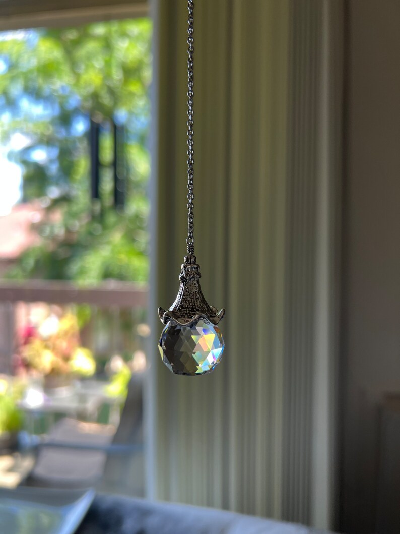 Op de afbeelding: Een gefacetteerde kristallen bol hanger hangt aan een zilveren ketting. Het kristal reflecteert een regenboog aan kleuren. De hanger hangt voor een raam met een vage buitenweergave.