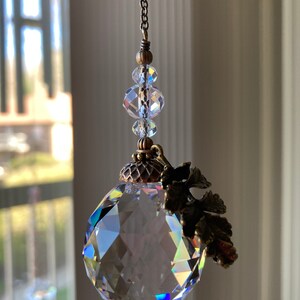 May include: A crystal suncatcher with a faceted ball and a bronze leaf design. The suncatcher is hanging from a chain with clear crystal beads.