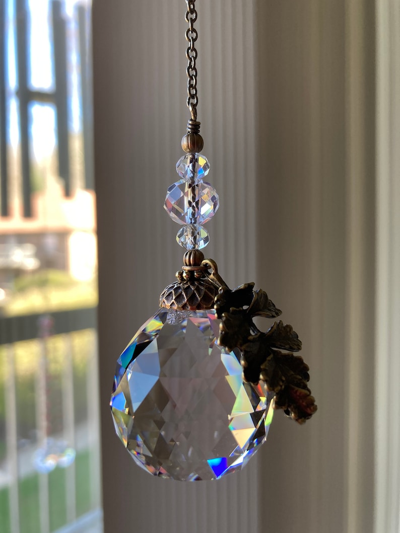 May include: A crystal suncatcher with a faceted ball and a bronze leaf design. The suncatcher is hanging from a chain with clear crystal beads.