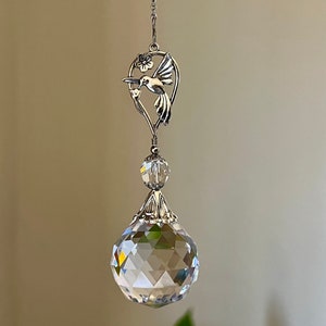 May include: A silver hummingbird with a crystal ball hanging from a chain. The hummingbird is perched on a branch with a flower. The crystal ball is clear and faceted.