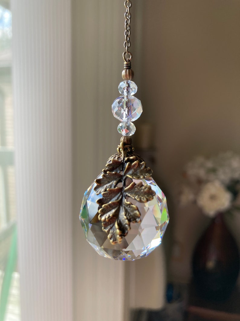 May include: A crystal suncatcher with a gold leaf design hangs from a chain with clear crystal beads.