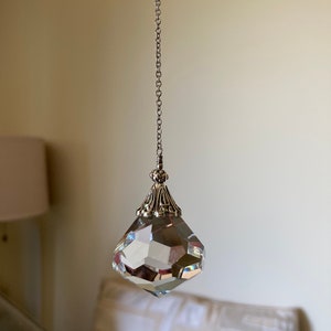 May include: A clear, faceted crystal prism pendant hanging from a silver chain. The prism has a decorative silver cap. The background is a neutral wall with a blurred lamp and pillow.