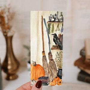 Peut inclure: Une peinture à l'aquarelle d'un atelier de sorcière avec deux balais, un corbeau noir, des citrouilles et des livres. La peinture est sur un fond blanc.